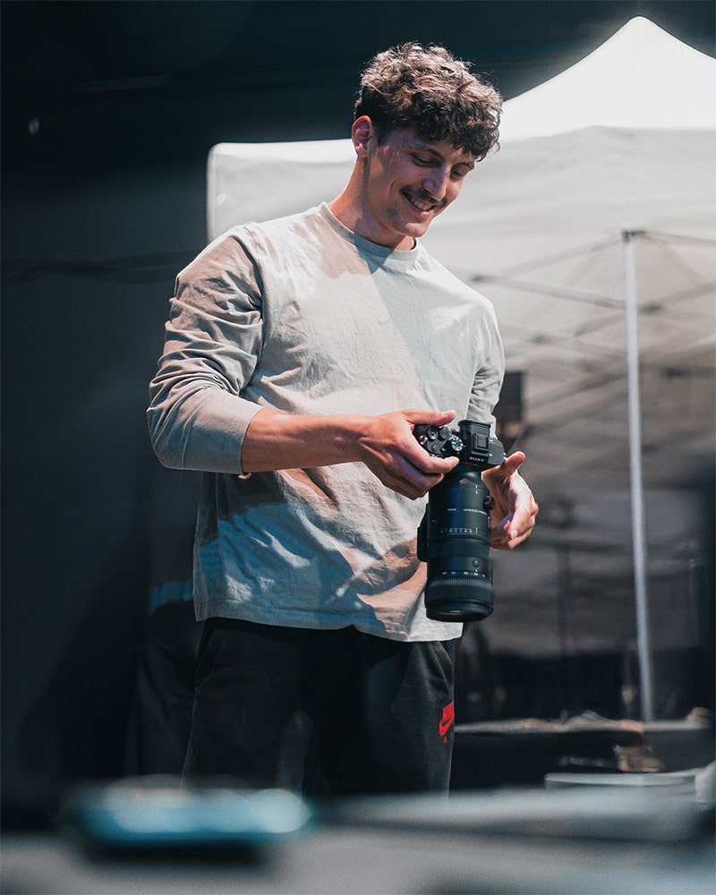 Photographe souriant en train de manipuler une caméra, habillé de manière décontractée avec un fond flou en arrière-plan.